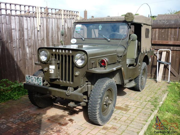 an old army truck parked in front of a wooden fence and brick walkway with grass growing on the ground