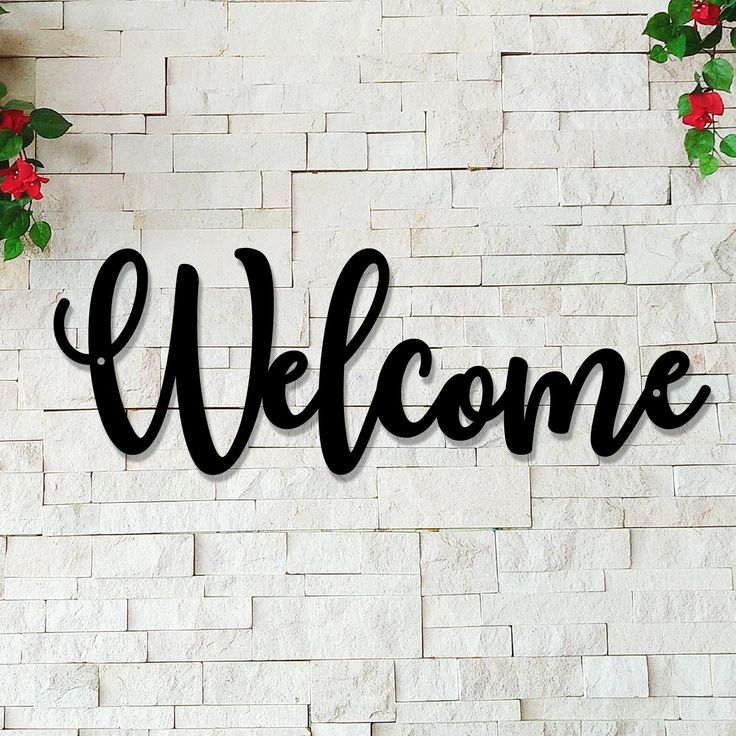 a welcome sign on the side of a white brick wall with red flowers hanging from it