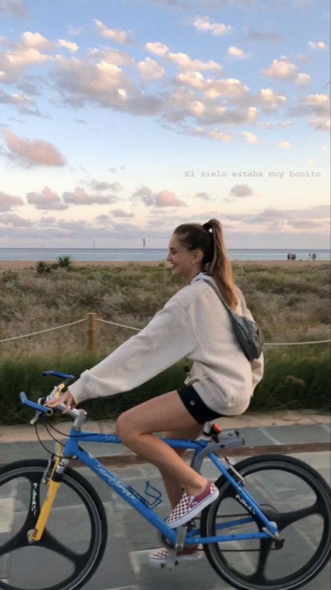 a woman riding a blue bike down a street next to a field with grass and clouds