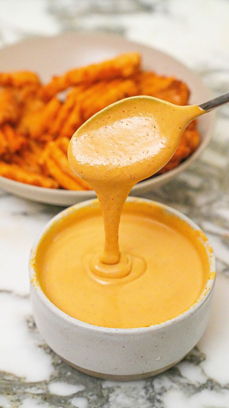 a spoon full of dipping sauce next to some sweet potato fries on a marble table