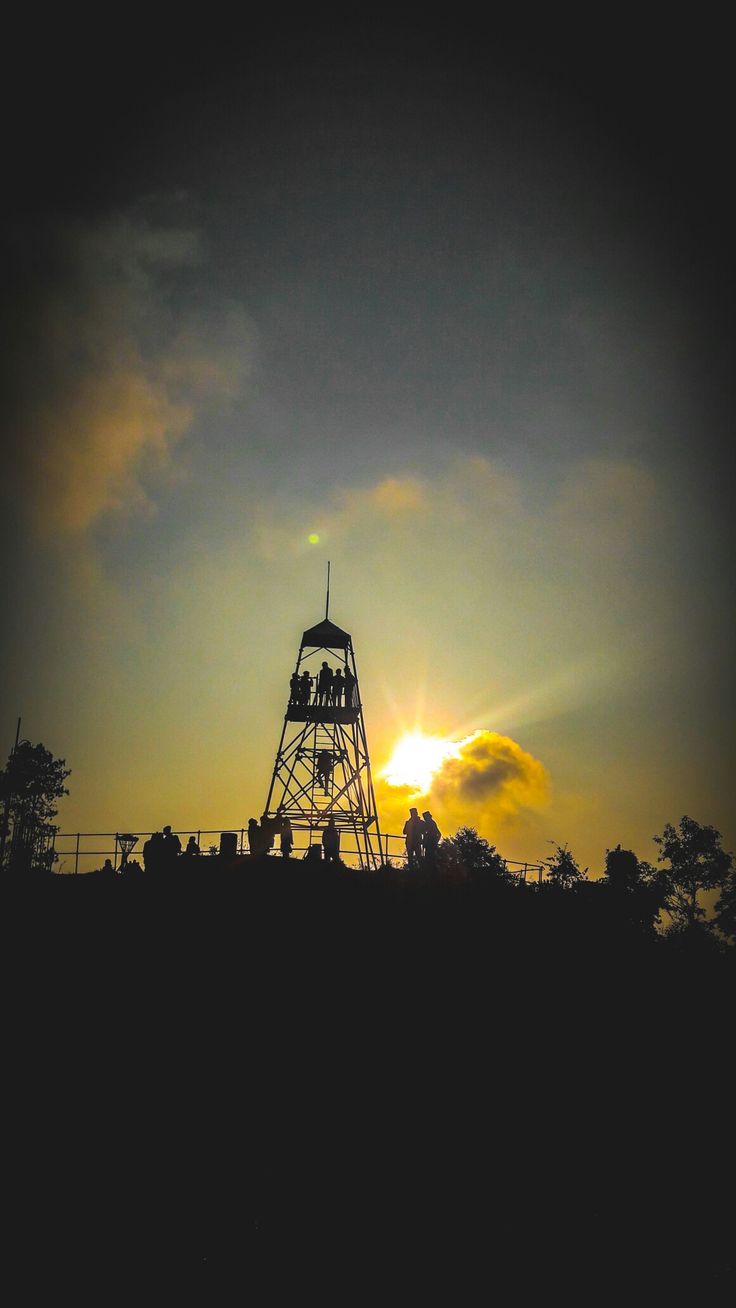 the sun is setting behind a tower on top of a hill with people sitting on it