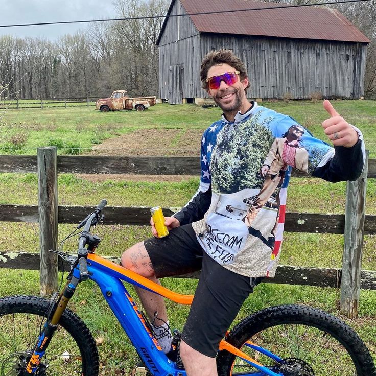 a man sitting on top of a blue bike in front of a fence with his thumbs up