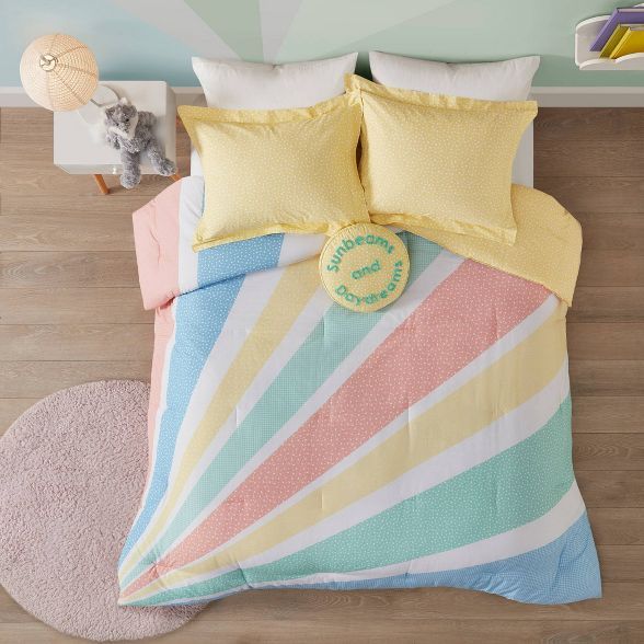 a bed with yellow, blue, and pink comforter on top of it next to a teddy bear