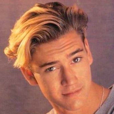 a man with blonde hair and blue eyes looks at the camera while wearing a gray shirt