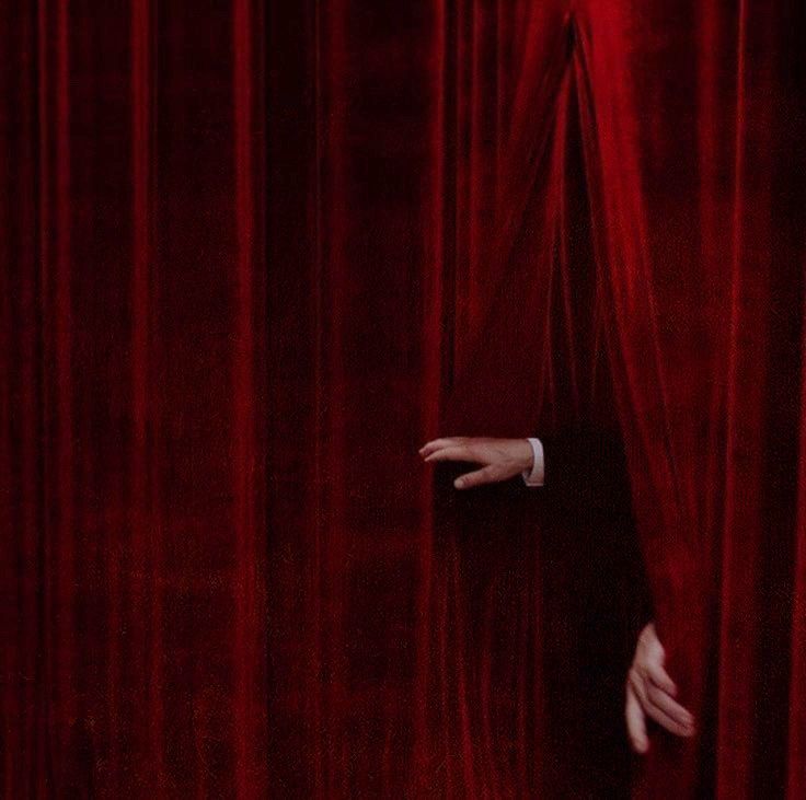 a man peeking out from behind a red curtain with his hand on the curtain edge