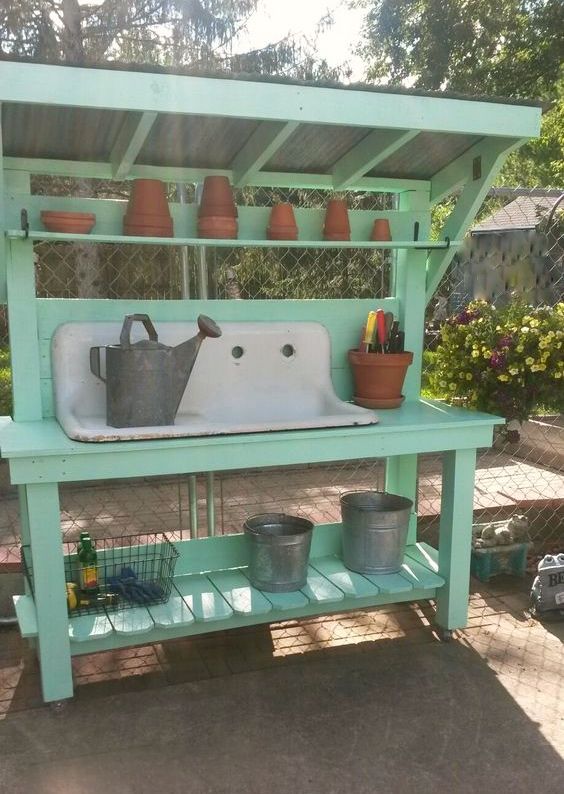 an outdoor sink with potted plants on the top and shelves above it, in front of a chain link fence