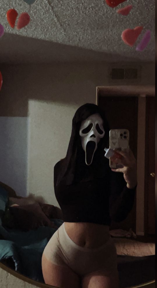 a woman wearing a ghost mask taking a selfie with her cell phone in the mirror