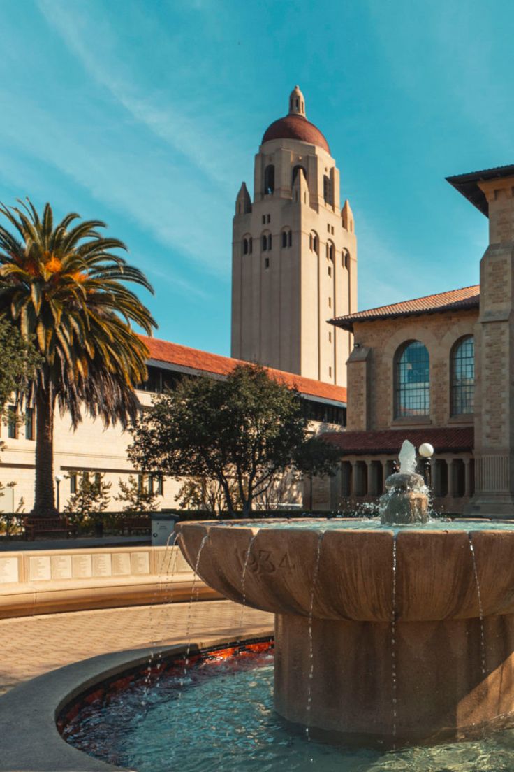 a fountain in front of a building with a clock tower on the top and palm trees around it