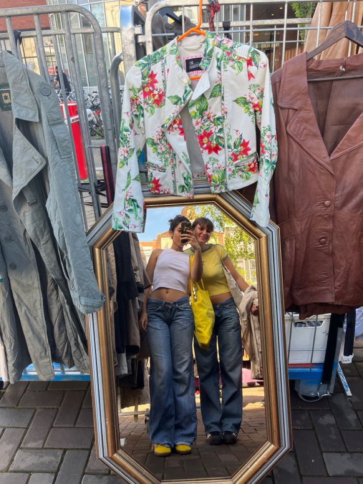 two women standing in front of a mirror with clothes hanging on the rack behind them