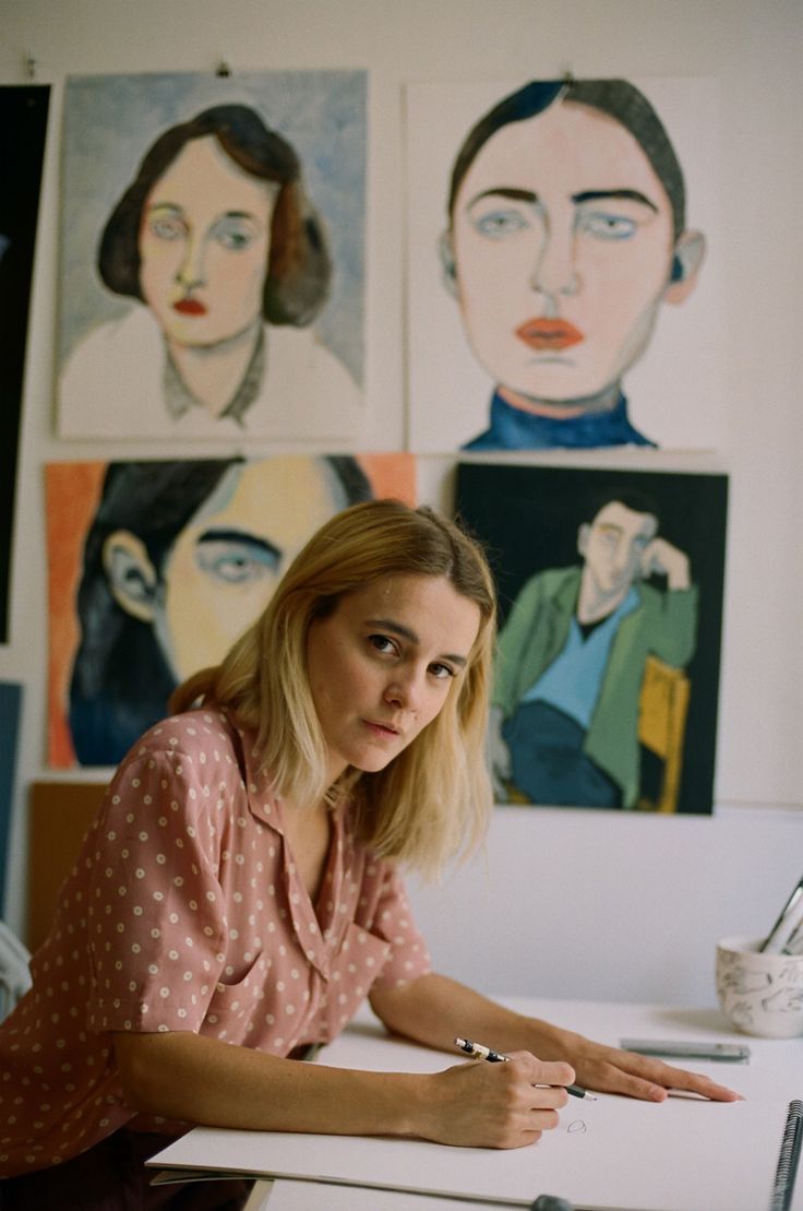 a woman is sitting at a desk in front of some paintings and pointing to the side