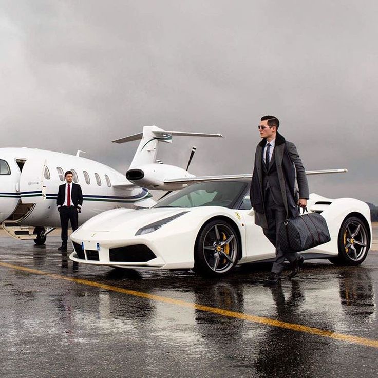 two men in suits standing next to a white sports car with a private jet behind them