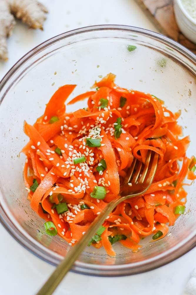 a glass bowl filled with carrots and topped with sesame seeds, garnished with green onions