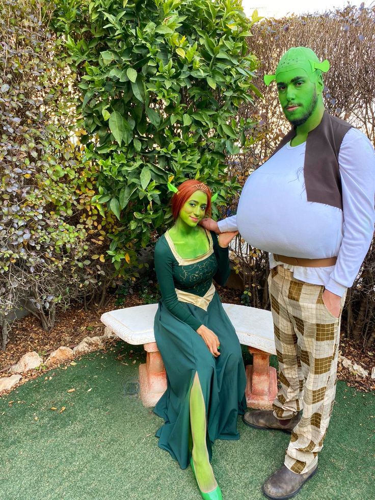 a man and woman dressed up as the same person sitting on a bench in front of bushes