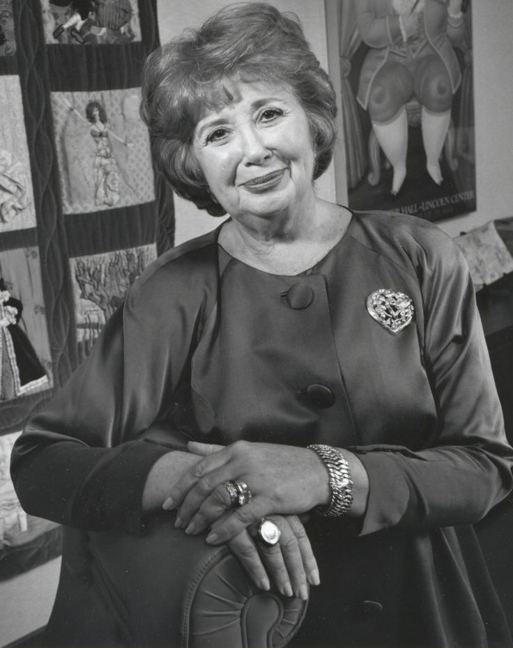 an older woman sitting in front of a wall with pictures on the walls behind her