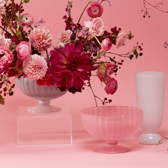 pink flowers are in a white vase next to other glass objects on a pink background