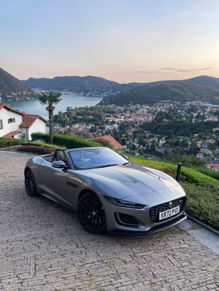 a grey sports car parked on top of a brick road next to a lush green hillside