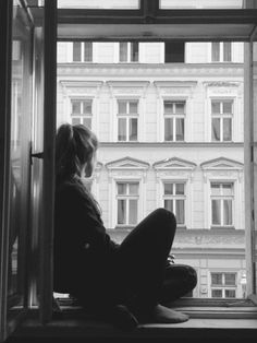 black and white photograph of woman sitting on window sill looking out at building across the street