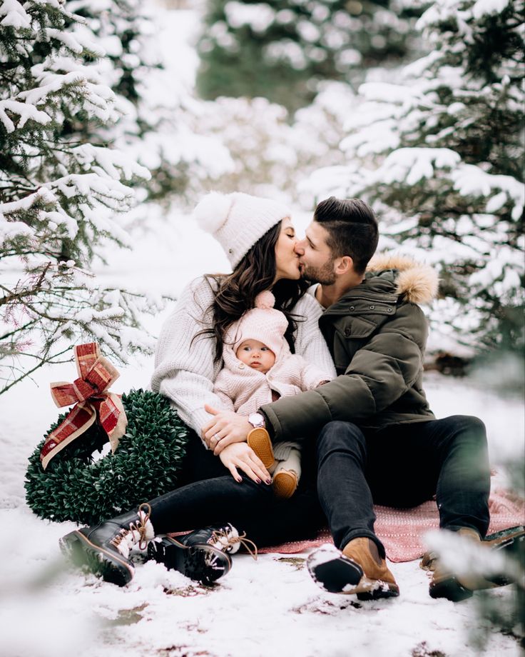 a man and woman kissing while sitting on a blanket in the snow next to trees