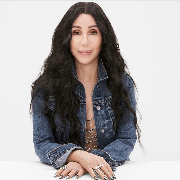 a woman with long black hair sitting on top of a white table wearing a jean jacket