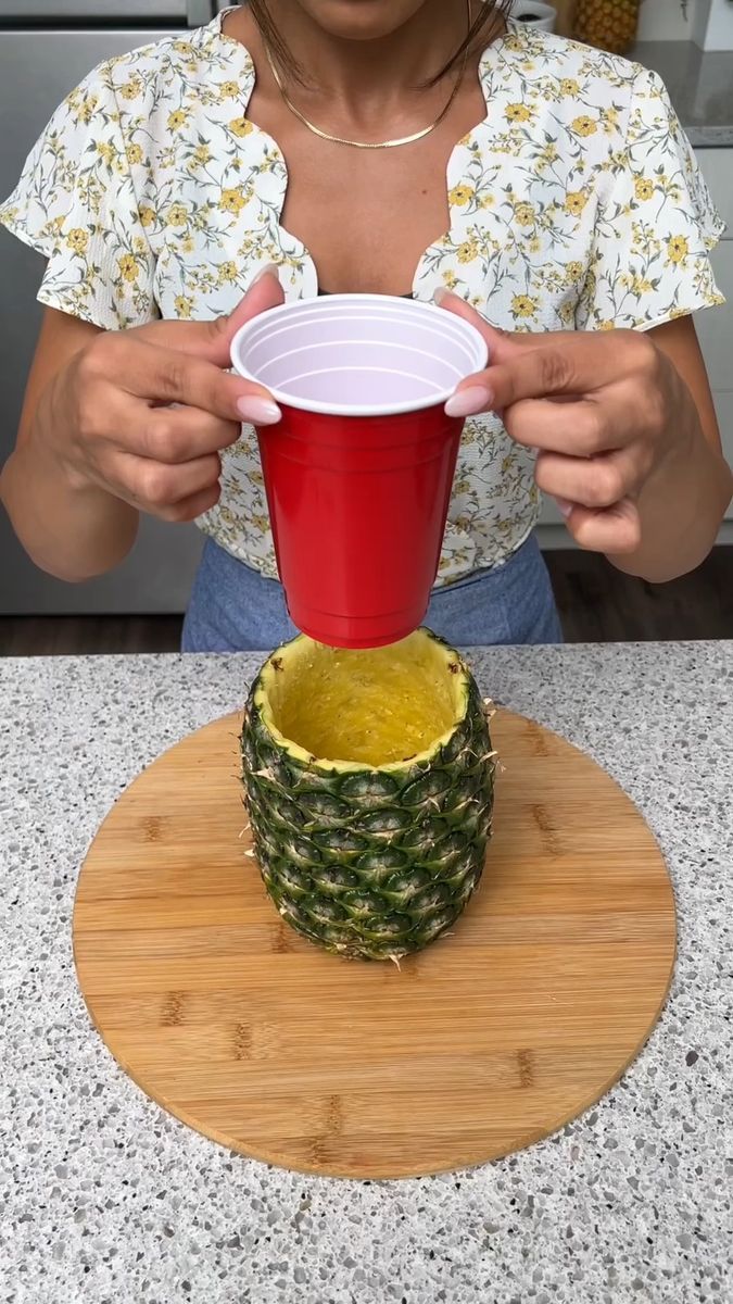 a woman holding a red cup on top of a wooden cutting board next to a pineapple