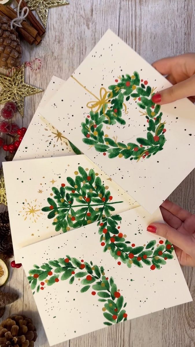 three handmade christmas cards with holly wreaths on them and pine cones in the background