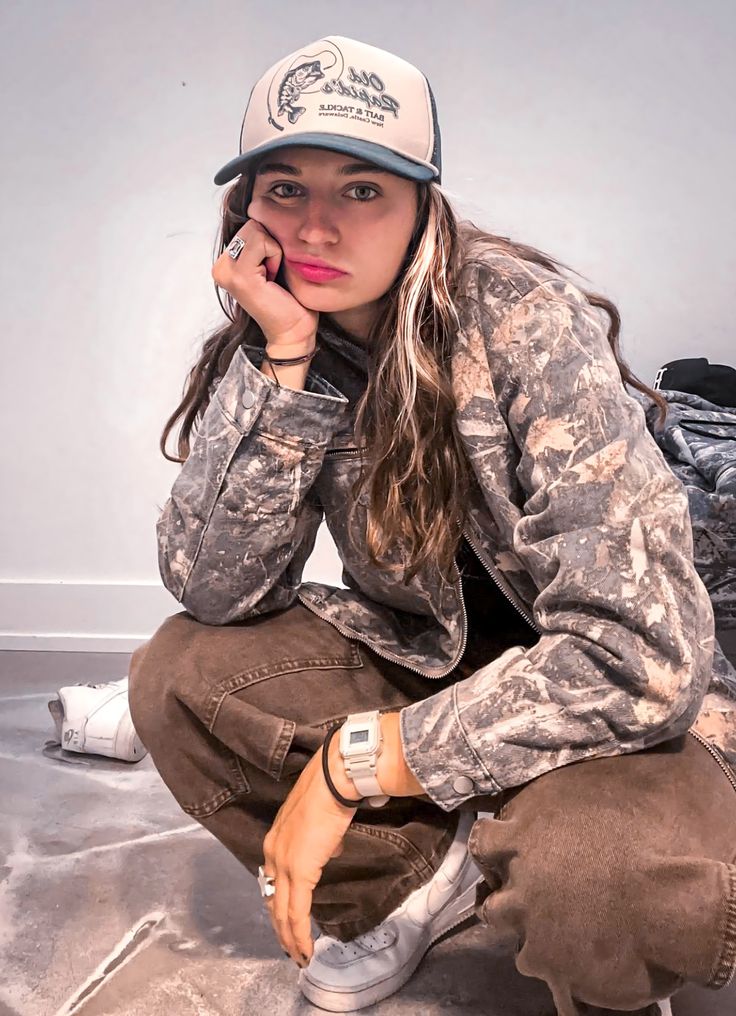 a woman in camo jacket and hat sitting on the ground with her hand under her chin