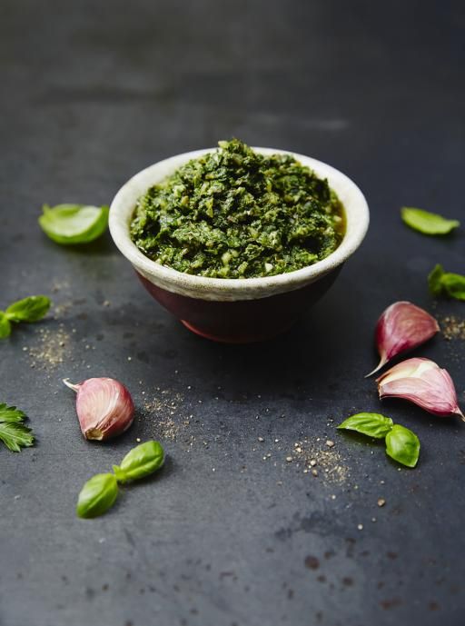 a small bowl filled with green pesto next to garlic and herbs on a table