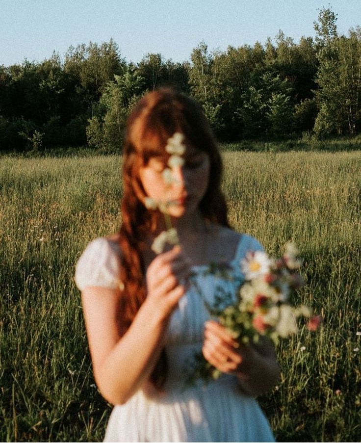 a girl in a white dress holding flowers and looking at the camera with trees in the background