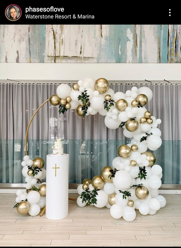 the balloon arch is decorated with gold and white balloons, greenery, and a cross