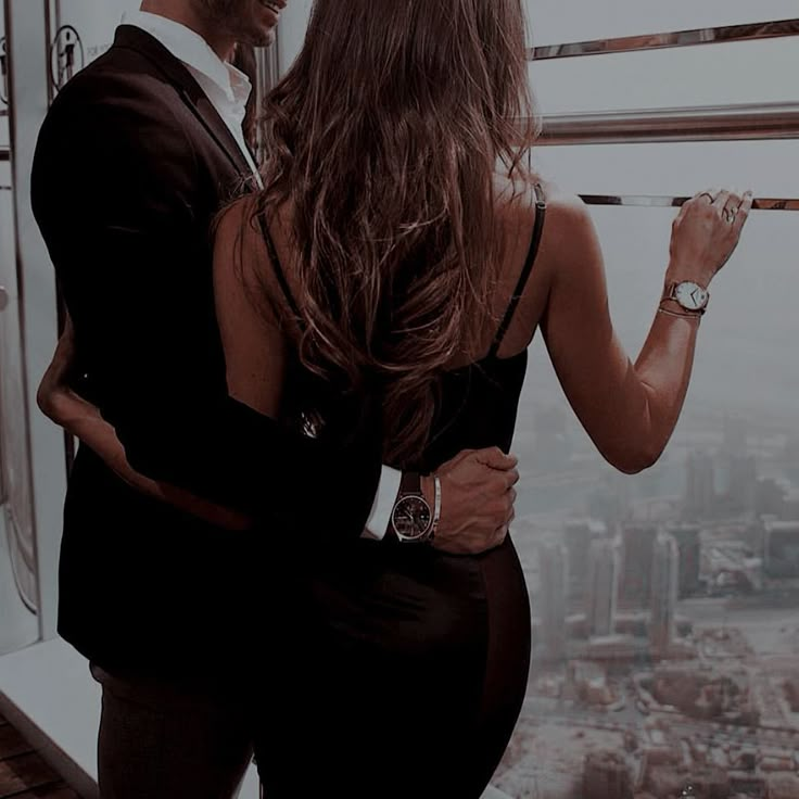 a man and woman standing next to each other in front of a window looking at the city