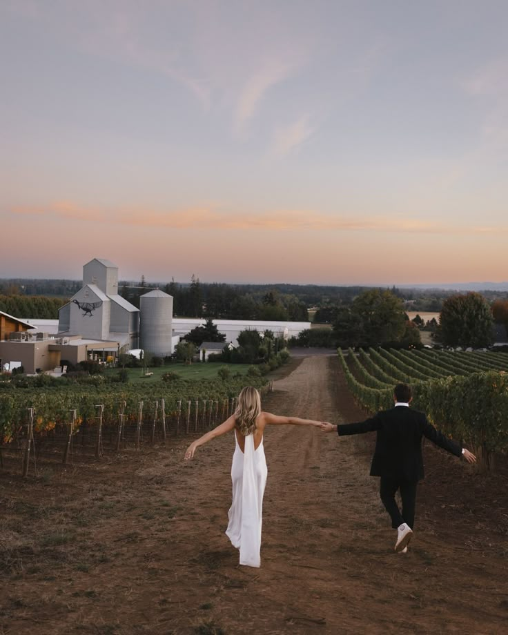 a man and woman holding hands walking down a dirt road in front of a vineyard