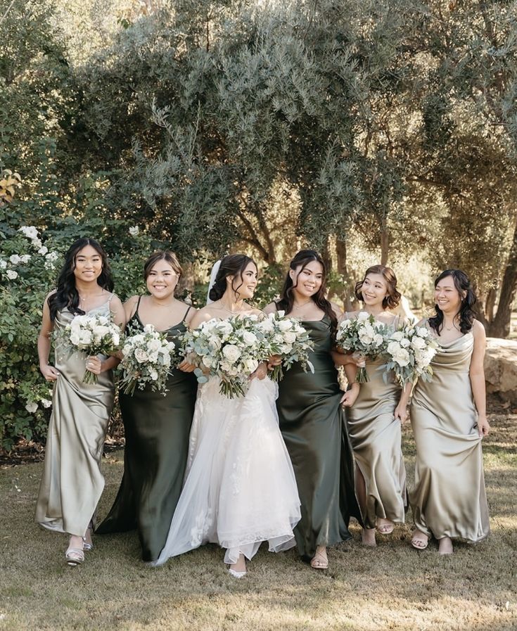 a group of women standing next to each other in front of trees and bushes holding bouquets