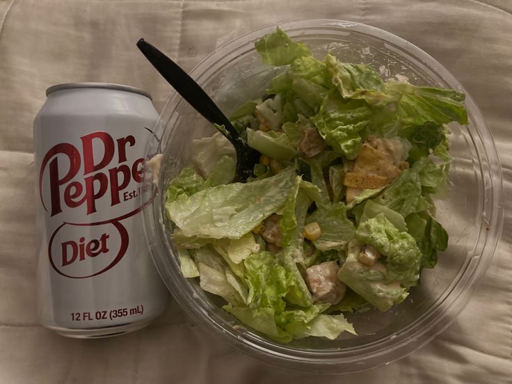 a salad in a plastic bowl next to a diet coke can on a white sheet