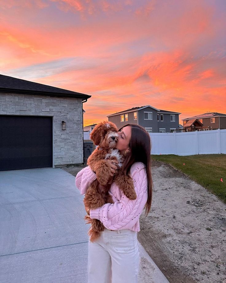 a woman holding a dog in her arms while the sun is setting behind her house