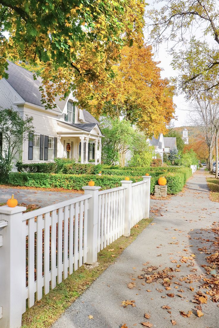 a white picket fence in front of a house with autumn leaves on the ground and trees