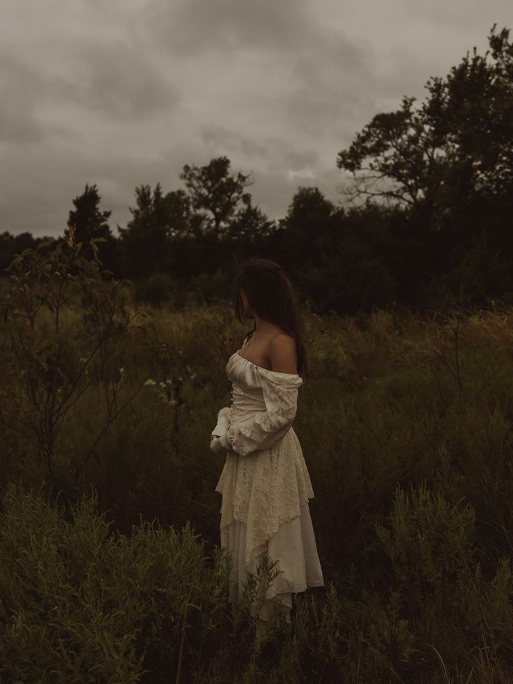 a woman in a white dress is standing in tall grass and looking off into the distance