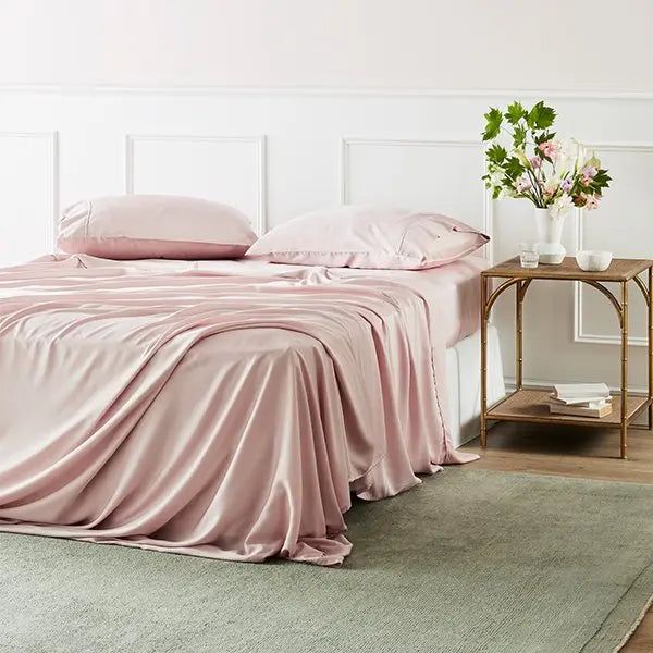 a bed with pink sheets and pillows in a white room next to a small table