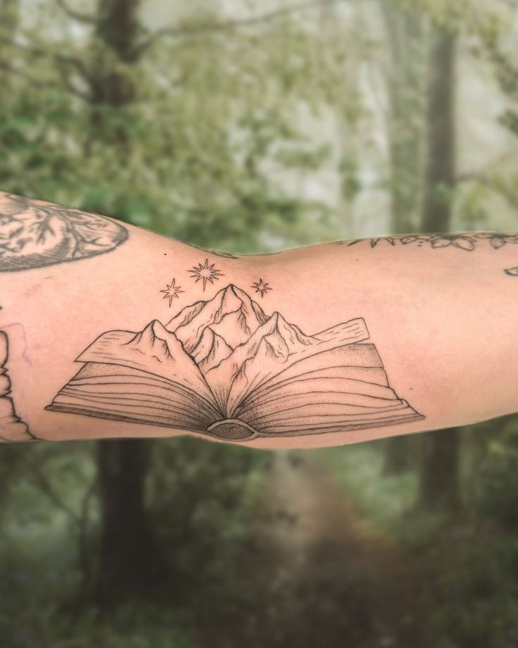 a man's arm with an open book tattoo on it and mountains in the background