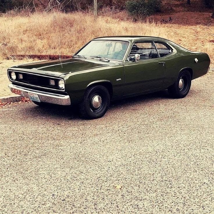 an old green car is parked on the side of the road in front of some dry grass