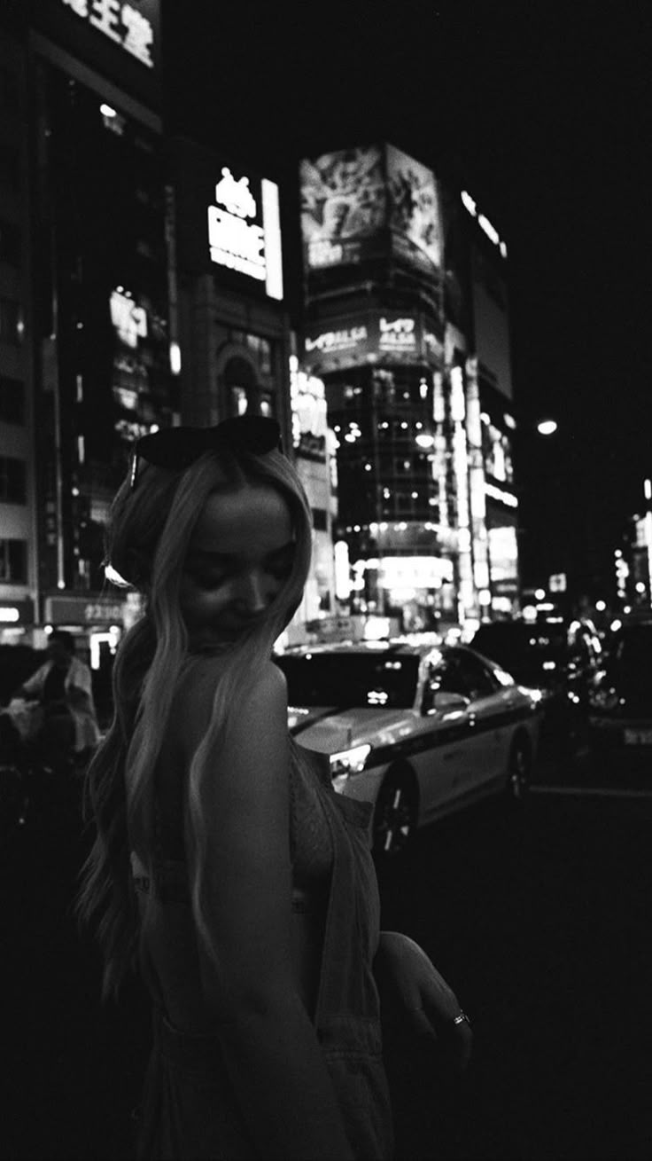 a black and white photo of a woman standing in the street at night with city lights behind her