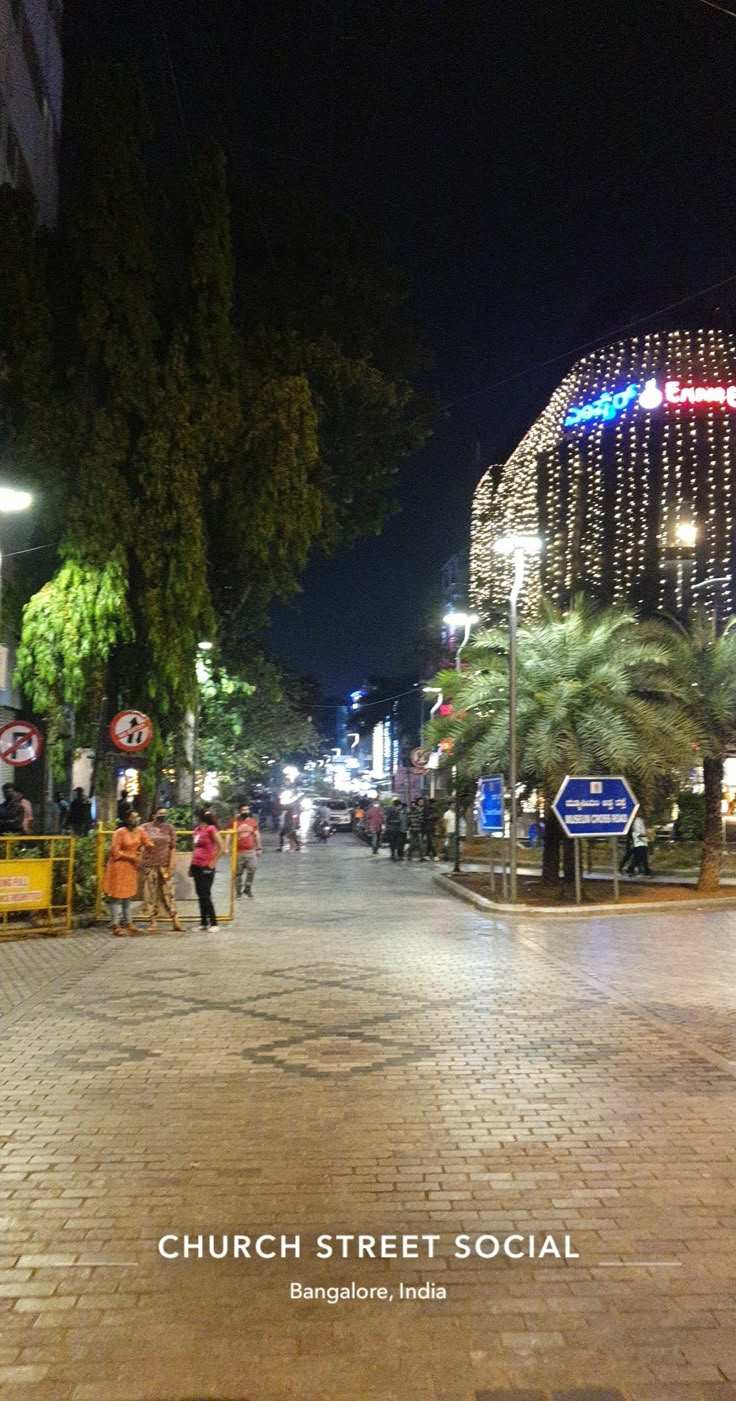 people walking down the street at night with lights strung up on buildings and palm trees