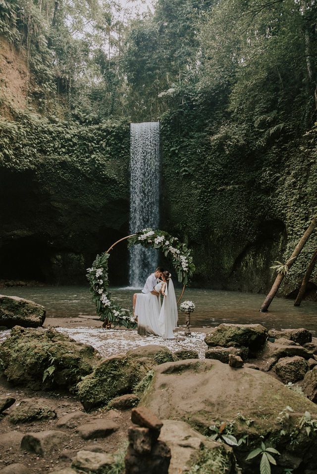 a bride and groom standing in front of a waterfall surrounded by greenery with flowers