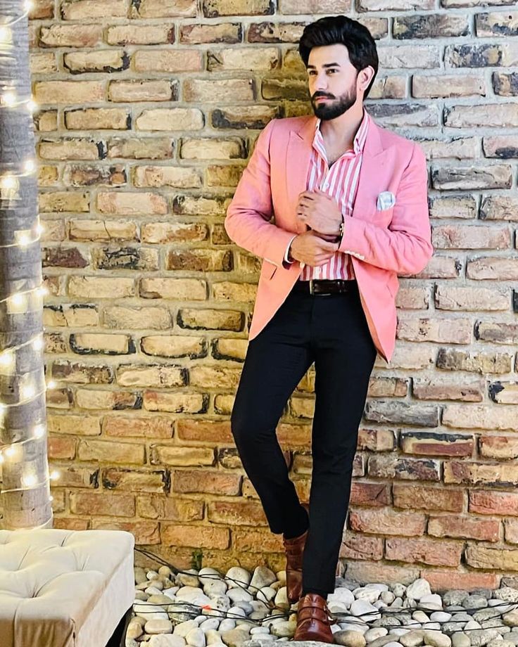 a man standing in front of a brick wall wearing a pink blazer and black pants