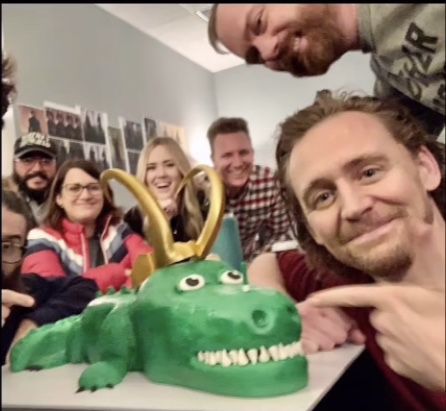 a group of people standing around a cake with a dinosaur on it's face