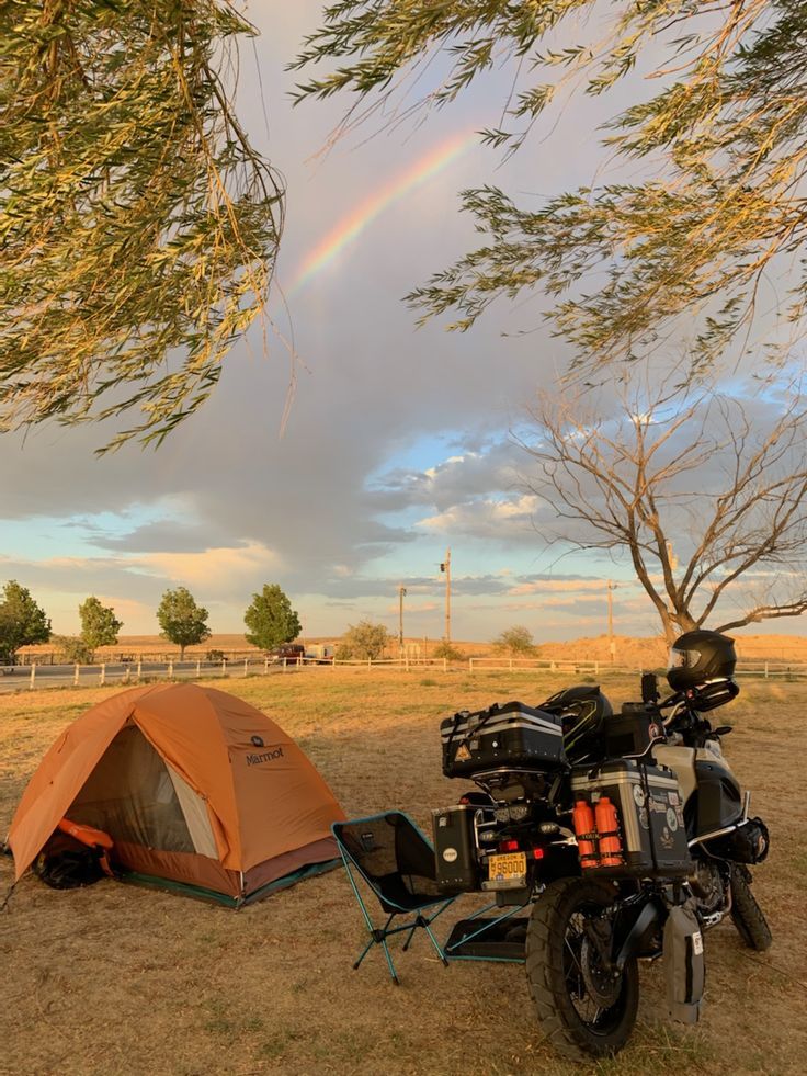 a motorcycle parked next to a tent under a tree with a rainbow in the background