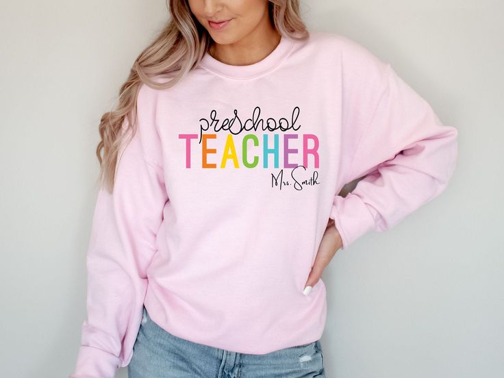 a woman wearing a pink sweatshirt with the words teacher on it, standing against a white wall