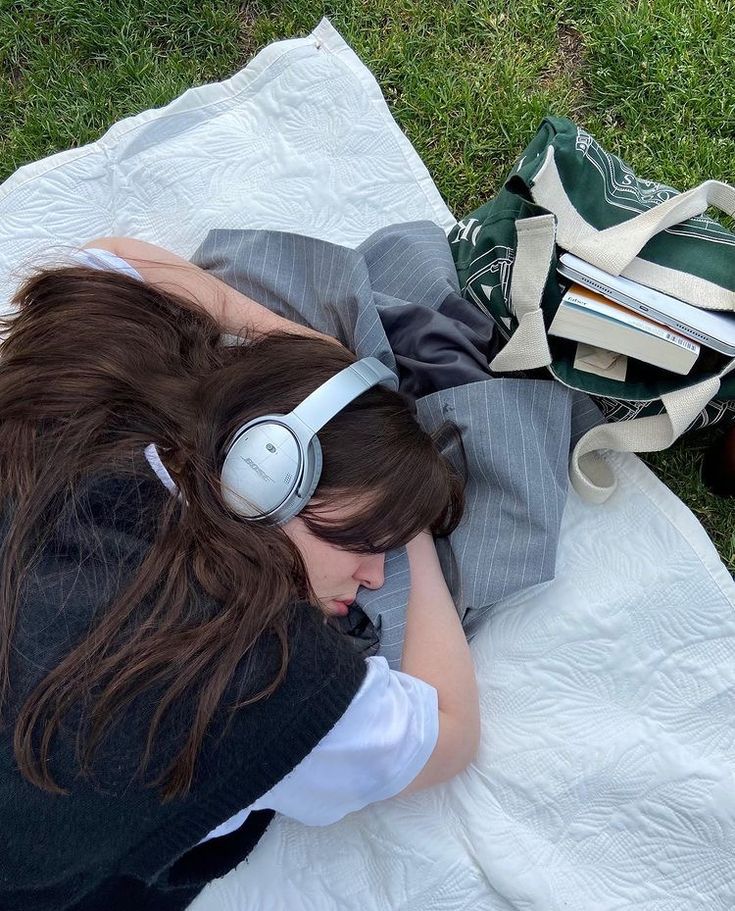 a woman laying on top of a blanket wearing headphones next to a purse and book bag