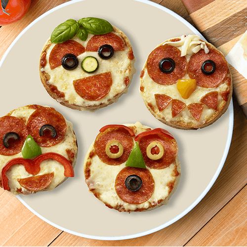 three small pizzas decorated like an owl on a table with vegetables and utensils