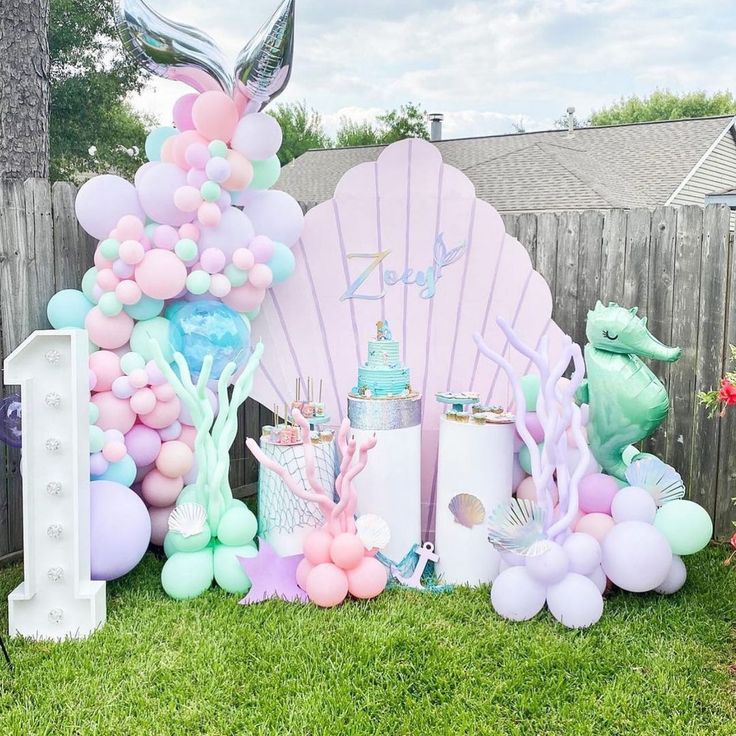 a mermaid themed birthday party with balloons, cake and other items on the grass in front of a fence