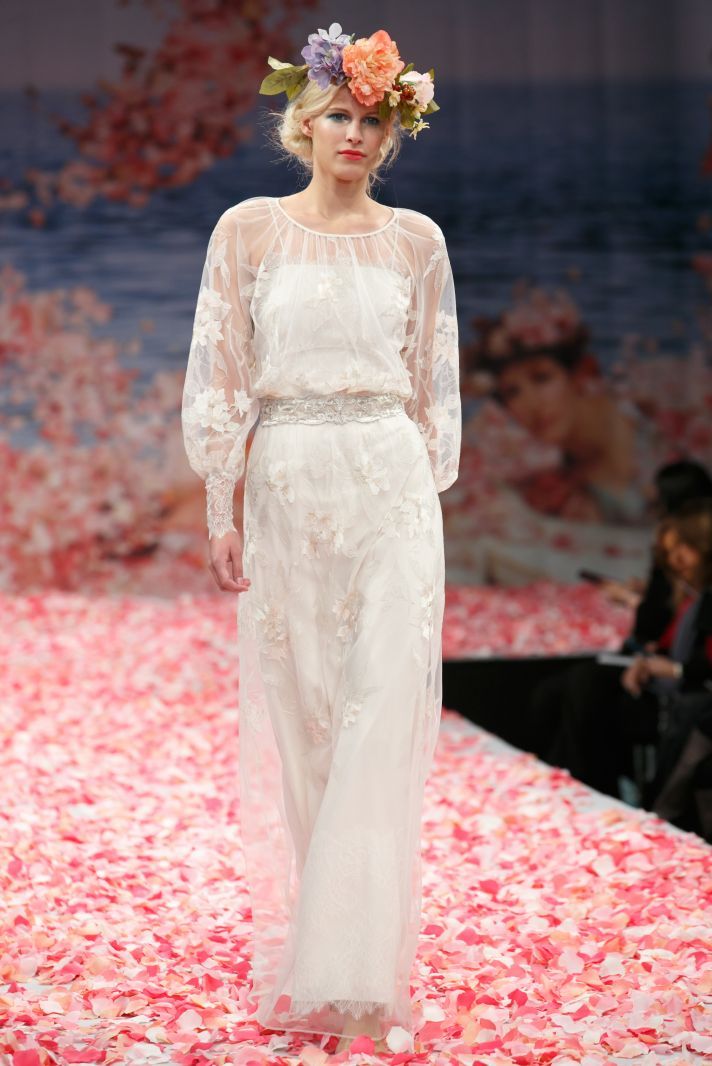 a woman walking down a runway wearing a white dress with flowers on her head and long sleeves